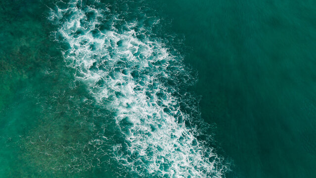Aerial top down view of ocean waves breaking over shallow coral reef creating white foam patterns in turquoise tropical water.