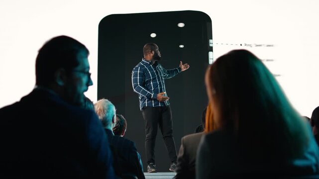 Black product manager introducing new mobile phone product at conference hall, featuring modern design and smart display. Launch event with audience, shareholders and potential clients. Camera A.