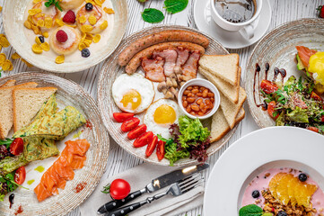 Breakfast spread at a cafe with various dishes and drinks served on white table. Several plates of breakfast food are placed on a white table with coffee and toast present as well.