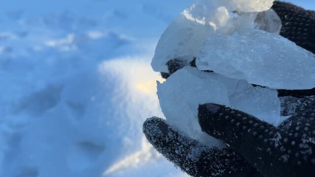 Holding a Large Chunk of Clear Ice. Black-gloved hands hold a large, jagged piece of clear ice against a snowy background, sunlight glinting through its thickness.