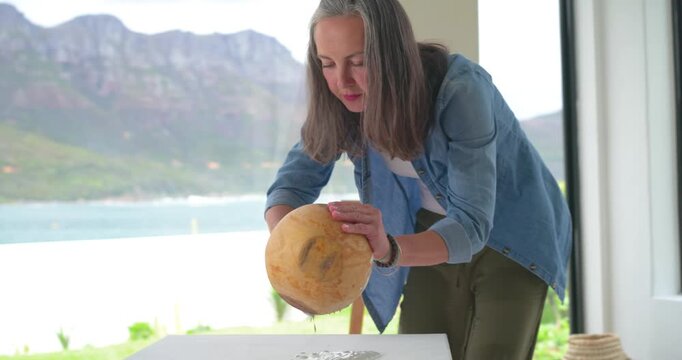 Mature adult woman inspecting underside, tilting gourd at small white table by bay window to drain