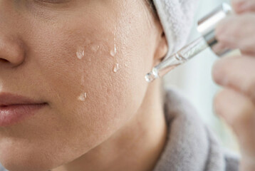 A close-up of skin care application. A hand holding a dropper placing drops on the cheek