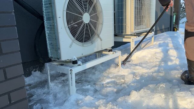 Shoveling Ice Chunks from Under Heat Pump. A black snow shovel removes a large pile of broken ice from beneath a heat pump, highlighting winter maintenance for air-source heating.