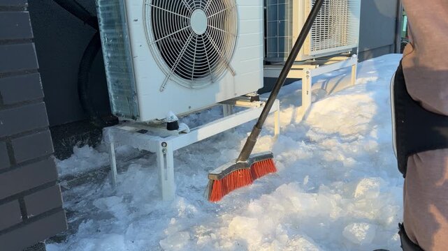 Clearing Ice from Under Heat Pump with Broom. A red broom sweeps shattered ice chunks from under a heat pump stand, maintaining airflow and drainage in winter conditions.