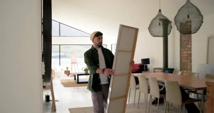 Testing canvas, Indian man in beanie lifting aligning canvas to hang on wall hook in hallway