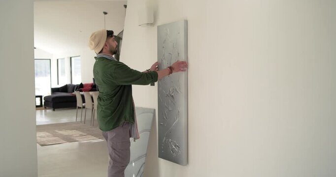 Indian man entering hall wearing beanie, removing mounted art and rehanging new canvas on wall hook