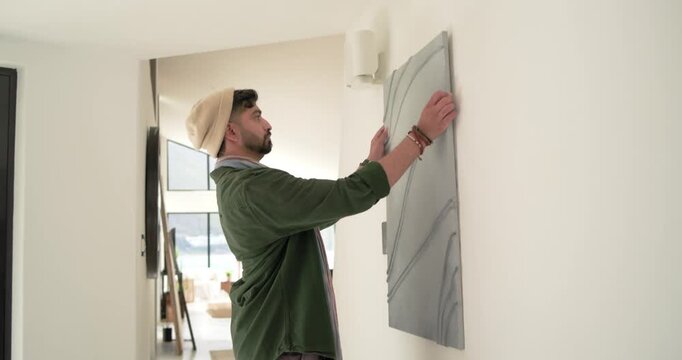 Adult Indian man in beanie noticing canvas, lifting, aligning to wall hook in hall, arranging decor