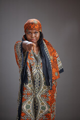 Obraz premium Young African woman wearing patterned traditional clothing and a headscarf points directly toward the camera while standing in a studio against a plain gray background.