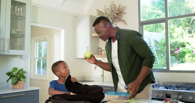 African American father and child packing school lunchbox and organizing backpack at kitchen island