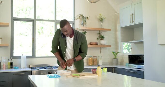 African American dad packing lunchbox at island, phone held, schoolboy arriving with blue backpack