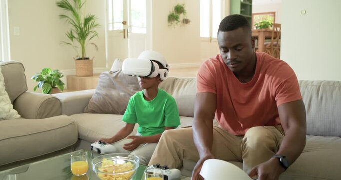African American father and child fitting white VR headset, handing controller, gaming on sofa