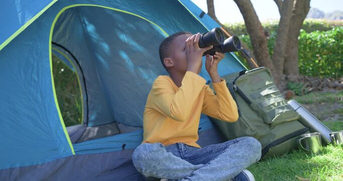 Opening African American boy sitting at blue tent raising binoculars, scanning sky by backpack