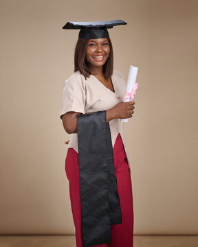 African woman wearing a graduation cap and black stole stands in profile, smiling while holding a rolled white certificate with a pink ribbon in a beige studio background setting.