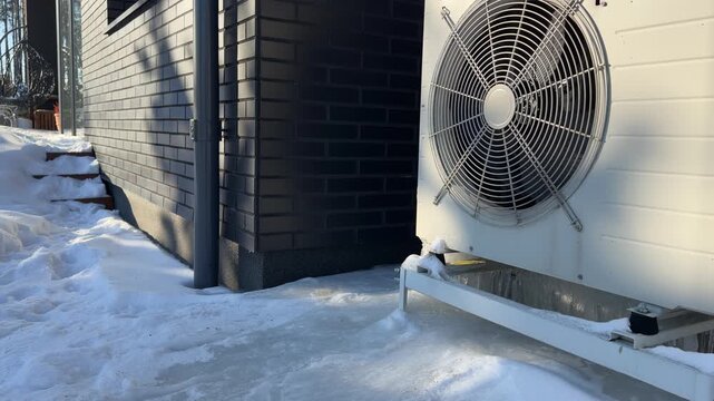 Heat Pump Unit Next to a Brick Wall in Winter. A white heat pump sits next to a dark brick wall surrounded by snow and ice, operating on a sunny, freezing winter day.