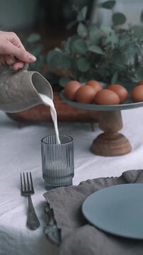 Pouring Fresh Milk from Ceramic Jug into Glass on Rustic Breakfast Table