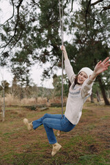 Young woman on a swing outdoors in a park, smiling and carefree, wearing a knit sweater and jeans, with trees and open space behind for a natural playful mood.