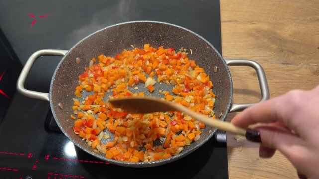 Cooking and Stirring Diced Vegetables in a Pan. A top-down view shows diced vegetables being stirred in a speckled frying pan on an induction cooktop, cooking evenly.
