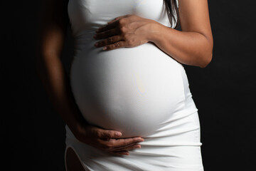 Close-up of a pregnant woman wearing a white dress, gently surrounding her baby bump with both hands. The face is not visible, keeping the focus on the belly and the protective gesture. Concept of pre