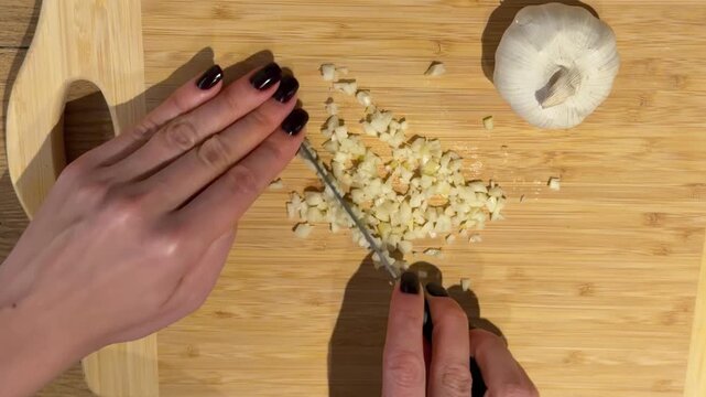 Finely Mincing Garlic on a Wooden Cutting Board. A top-down close-up captures hands finely mincing a pile of garlic on a wooden board with a knife, a whole bulb resting nearby.