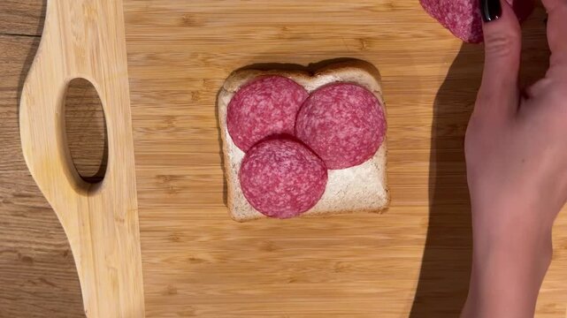 Placing Salami Slices on Buttered Toast. A top-down shot shows hands placing round salami slices onto buttered toast on a wooden board, assembling a classic sandwich.