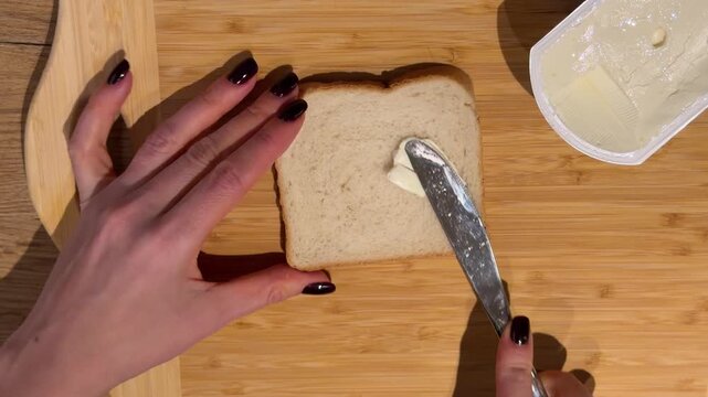 Buttering a Slice of White Bread for a Sandwich. A top-down view captures hands spreading a layer of butter on a slice of white bread on a wooden board, preparing for a sandwich.
