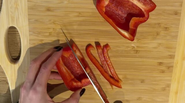 Slicing a Red Bell Pepper into Strips on a Wooden Board. A top-down view shows hands slicing a red bell pepper into thin strips on a wooden board, preparing fresh vegetables with a sharp knife.