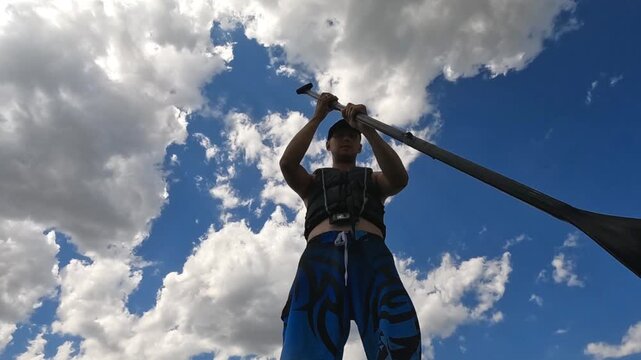 Low angle: a man in a life jacket rowing on a SUP board. Silhouette against the background of a bright sky with clouds. Sports, summer, freedom.