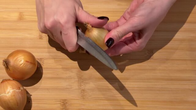 Peeling an Onion on a Wooden Cutting Board Top Down. A top-down view shows hands peeling the outer skin from a whole onion on a wooden board, capturing the initial step of food preparation.