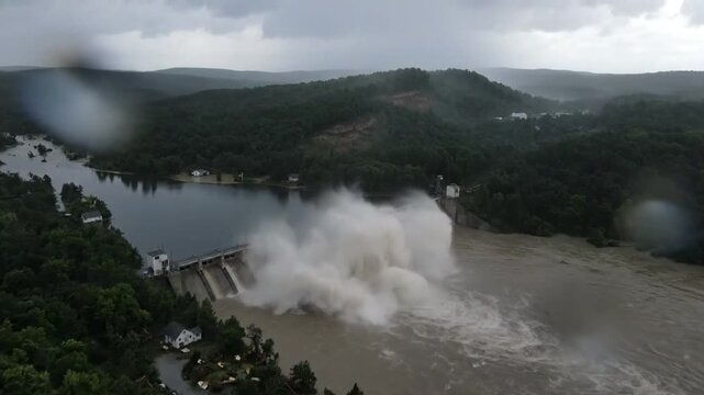 Drone view of a dam exploding and releasing a massive torrent of water into the river during heavy rainfall.