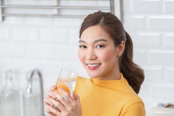 Beautiful young Asian woman in a yellow shirt looking at the camera and smiling while holding a glass of fresh orange-infused water with ice in a bright kitchen.