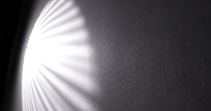 An abstract and captivating play of light and shadow dynamically interacts with a textured wall surface. Brilliant rays of light, fanned out in a striking pattern, slowly sweep across the dark, rugged