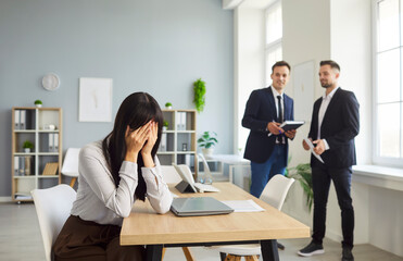 Upset female worker hides her face while two colleagues chat behind, hinting at gossip, bullying...