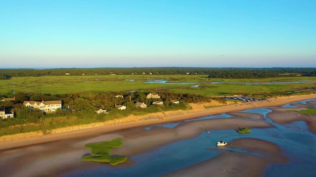 Drone sunset aerial of Cape Cod shoreline with tidal flats and shallow pools, beachgoers scattered along the sand, and green marshland. Scenic New England coast, smooth 4K b-roll.