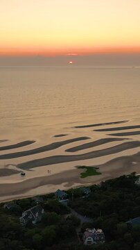 Vertical drone sunset over Cape Cod tidal flats, glowing reflection on calm water with braided sandbars and gentle surf. Tall-frame coastal seascape with soft clouds, cinematic stock b-roll.