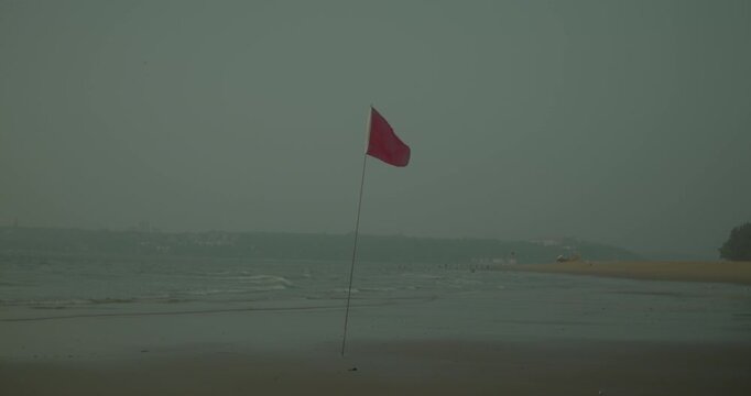 A red warning flag stands on a quiet beach as waves roll in under hazy, overcast conditions