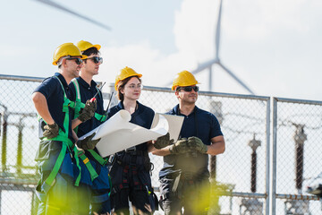 Professional group of diverse industrial engineers in safety gear and hardhats smiling and looking forward while holding technical plans and a laptop at an electrical substation.