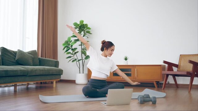 Happy young Asian woman practicing yoga and shoulder stretching at home sitting on floor in living room