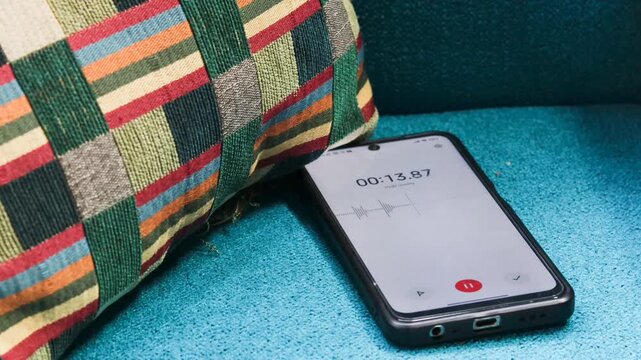 Voice detection and recording app in mobile phone listening to contestation laying on couch or sofa hidden from view. Secretly grabbing and wiretapping inside home or apartment during covert operation
