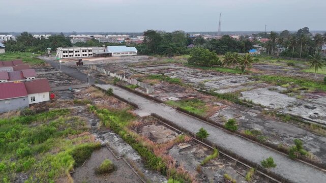 Aerial view of a large vacant land with concrete foundations, dirt roads, scattered vegetation, palm trees and surrounding residential houses at dusk.