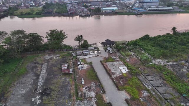 Aerial drone footage of vacant land on the riverbank featuring concrete foundations, dirt roads, scattered trees, and small huts along the wide Kapuas River.