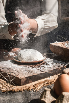 Close-up of professional baker hands clapping and dusting white flour powder over fresh bread dough on a wooden board. Frozen motion of flour particles in a dark, moody kitchen atmosphere. 