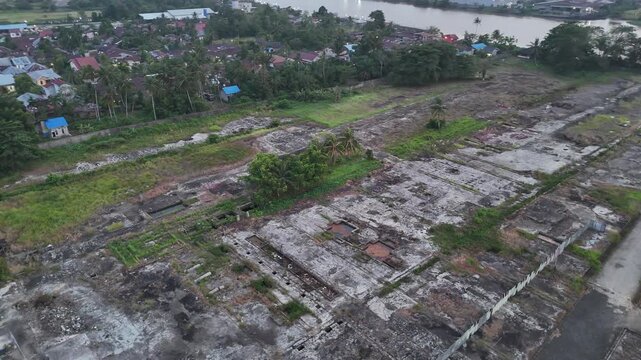 Aerial view of a large vacant riverside plot with cracked concrete foundations, scattered vegetation, palm trees, small structures and residential neighborhood near a wide river at dusk.