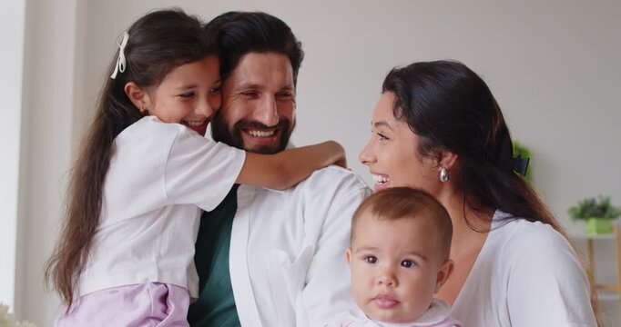 Happy family hugs at home, cheerful carefree parents holding kids in arms with love, care and support. Smiling father and mother talking, standing in living room, hugging toddler and teenage girl