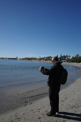 A man stands by the sea with a camera, ready to take a snapshot of the landscape on a sunny day.