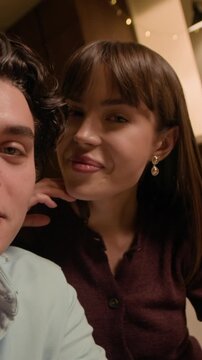 Vertical POV shot of young Caucasian man and woman talking on camera while vlogging in living room and drinking wine