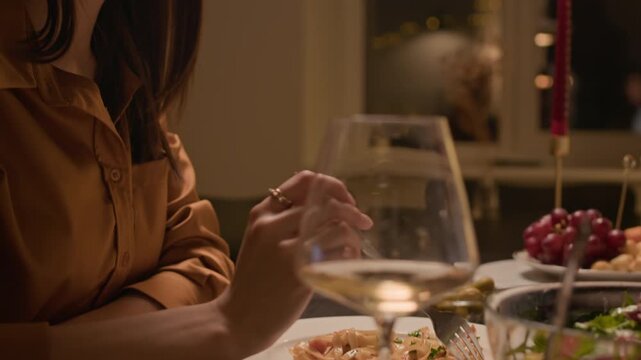Tilt up shot of young Caucasian lady eating pasta and drinking white wine while dining at festive table at home in warm light