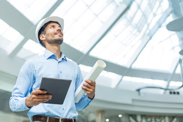 Fototapeta premium Young architect in white hard hat with plans looking up in a bright office in building under construction