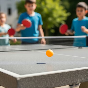 A table tennis ball bouncing on an outdoor stone ping pong table, with children playing in the blurred background, highlighting youthful recreation.