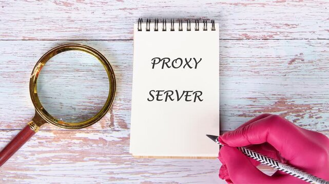 A gloved hand writes Proxy Server on a small spiral notepad with a pen. A magnifying glass rests on the light wooden desk, emphasizing the focus on this computing term