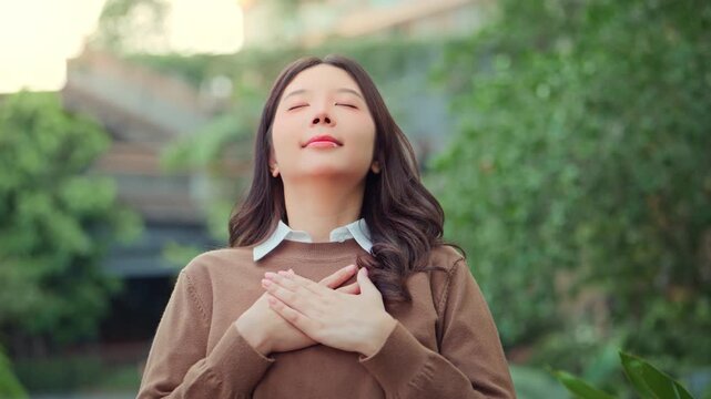 Peaceful Asian woman closing eyes with hands on chest expressing gratitude mindfulness and inner peace. Mental health wellness meditation self-care and emotional wellbeing concept.
